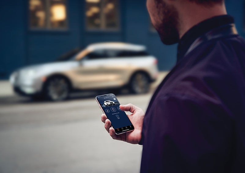 A person is shown interacting with a smartphone to connect to a Lincoln vehicle across the street. | Libertyville Lincoln Sales, Inc. in Libertyville IL