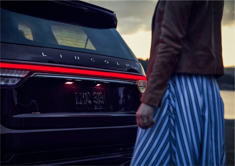 A person is shown near the rear of a 2023 Lincoln Aviator® SUV as the Lincoln Embrace illuminates the rear lights | Libertyville Lincoln Sales, Inc. in Libertyville IL