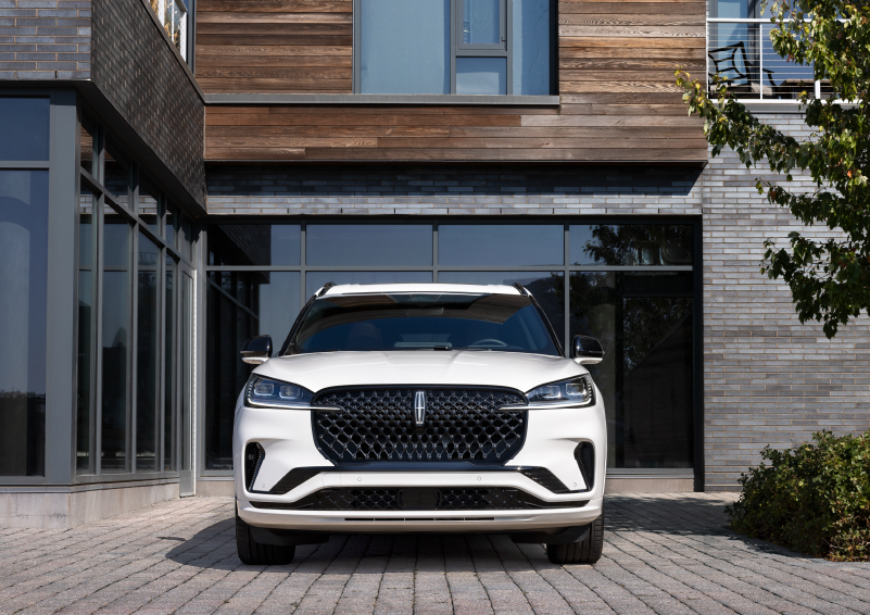 A white 2025 Lincoln Aviator® SUV is shown parked outside a modern building with gray brick and wood accents. | Libertyville Lincoln Sales, Inc. in Libertyville IL