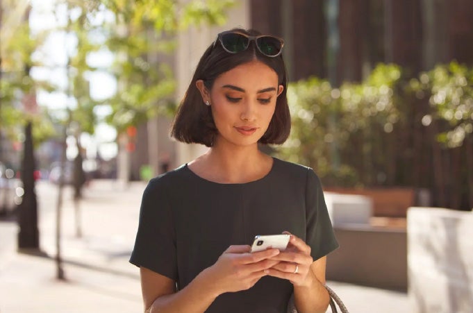 A woman looking at her phone on a sun-dappled city sidewalk.